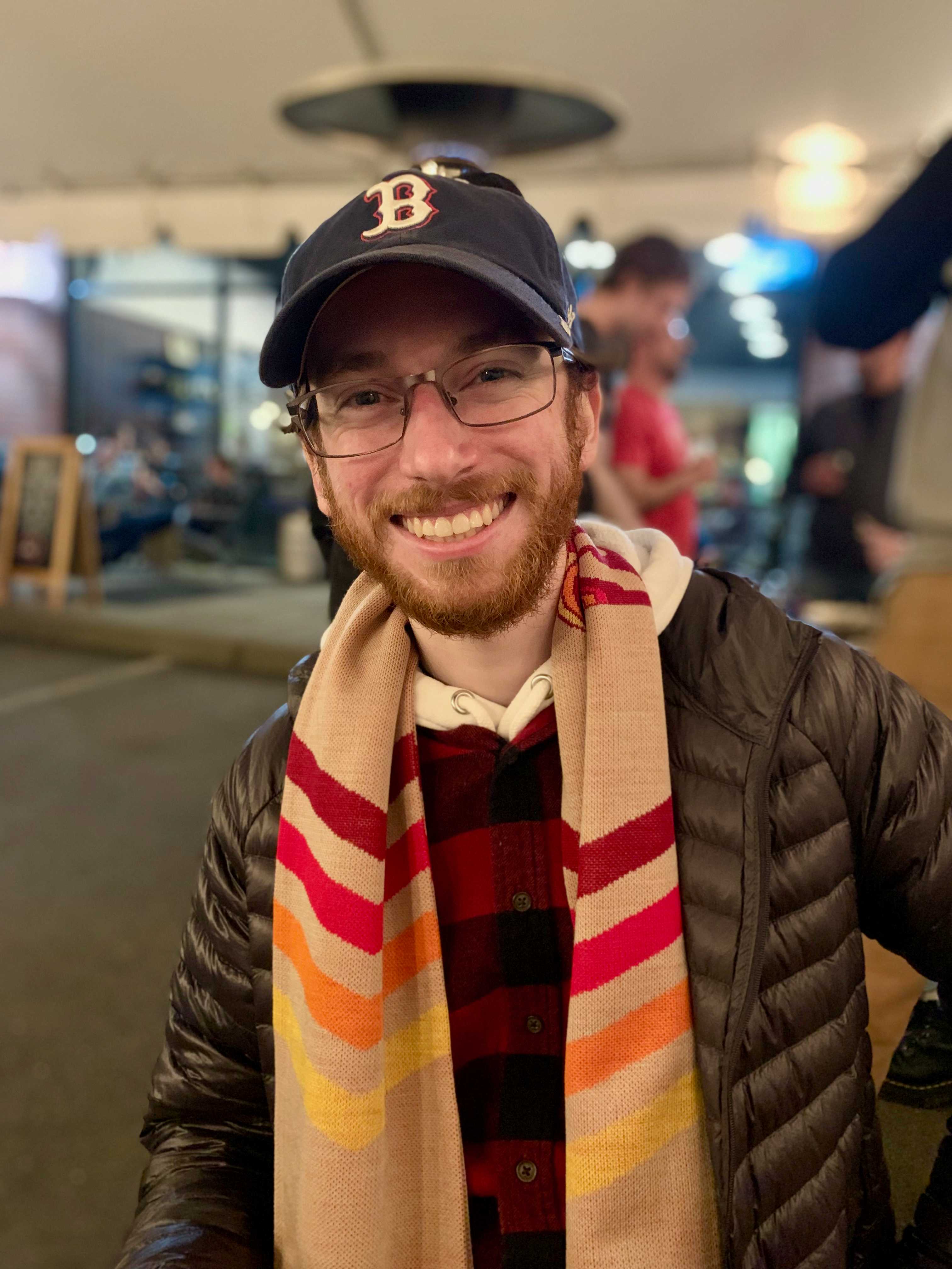 Sam wearing a Boston Red Sox baseball cap and a tan scarf with the Portland Thorns logo. Sam is a white man, in his late 20's with a short red-ish brown beard.